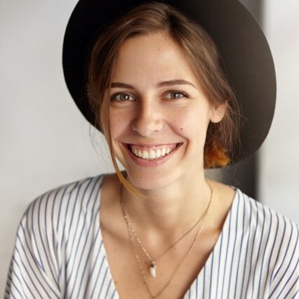 Fashionable cute young lady with dark eyes, healthy skin smiling demonstrating her white teeth wearing black hat being glad to have holidays. Woman with appealing appearance rejoicing her holidays